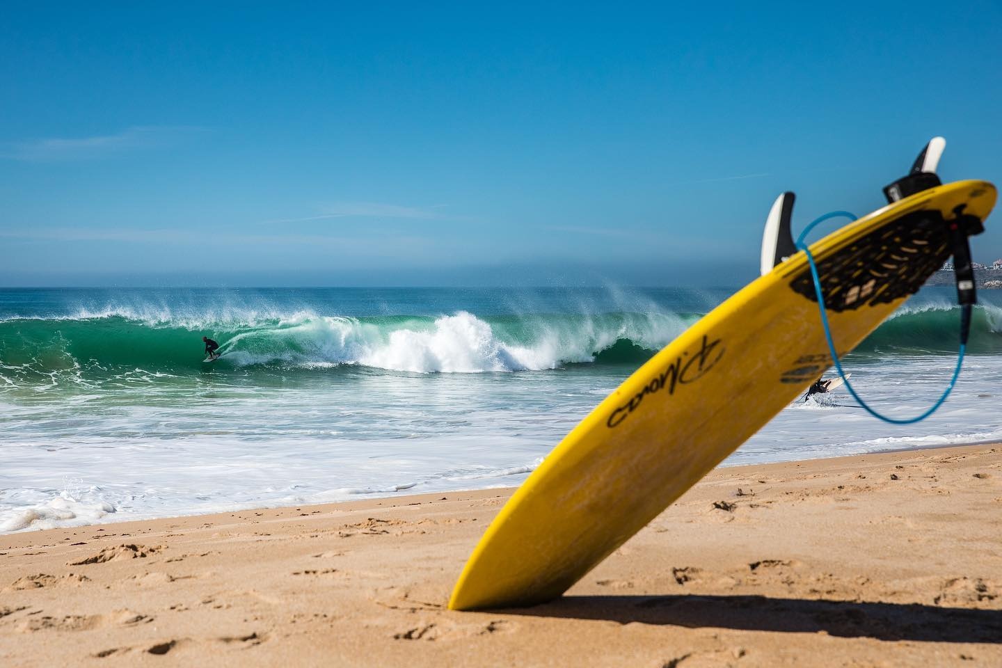Uma prancha de surf amarela está inclinada na areia da praia, com um surfista a apanhar uma grande onda verde no mar ao fundo.