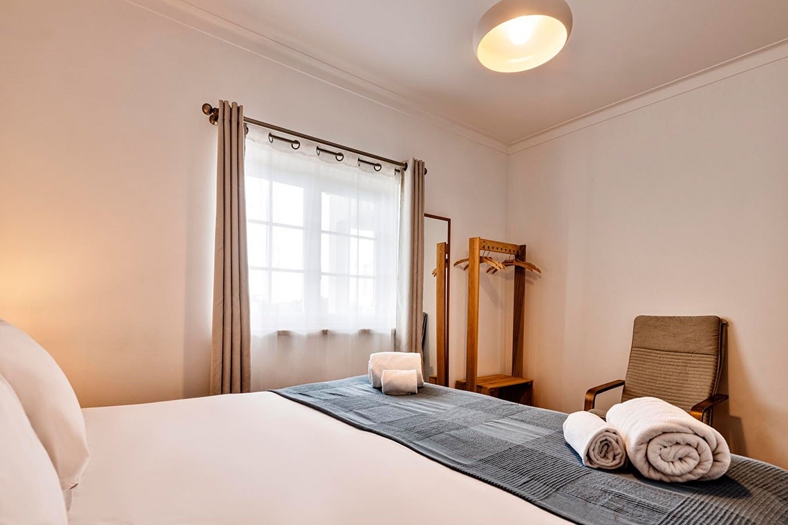 Um quarto de dormir claro e arrumado, com uma cama feita, cortinas na janela, um cabide de madeira e uma poltrona.