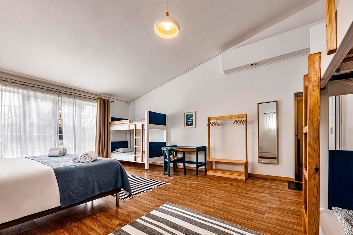 Um quarto amplo e luminoso, decorado com uma cama de casal, beliches, chão de madeira, uma secretária, um cabideiro e janelas grandes com cortinas.