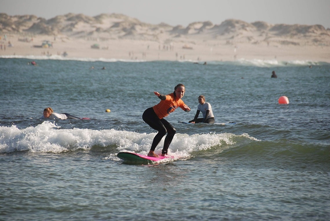 Uma mulher de camisa laranja e calças pretas surfa uma pequena onda em uma prancha rosa, com outras pessoas na água e uma praia arenosa com dunas ao fundo.