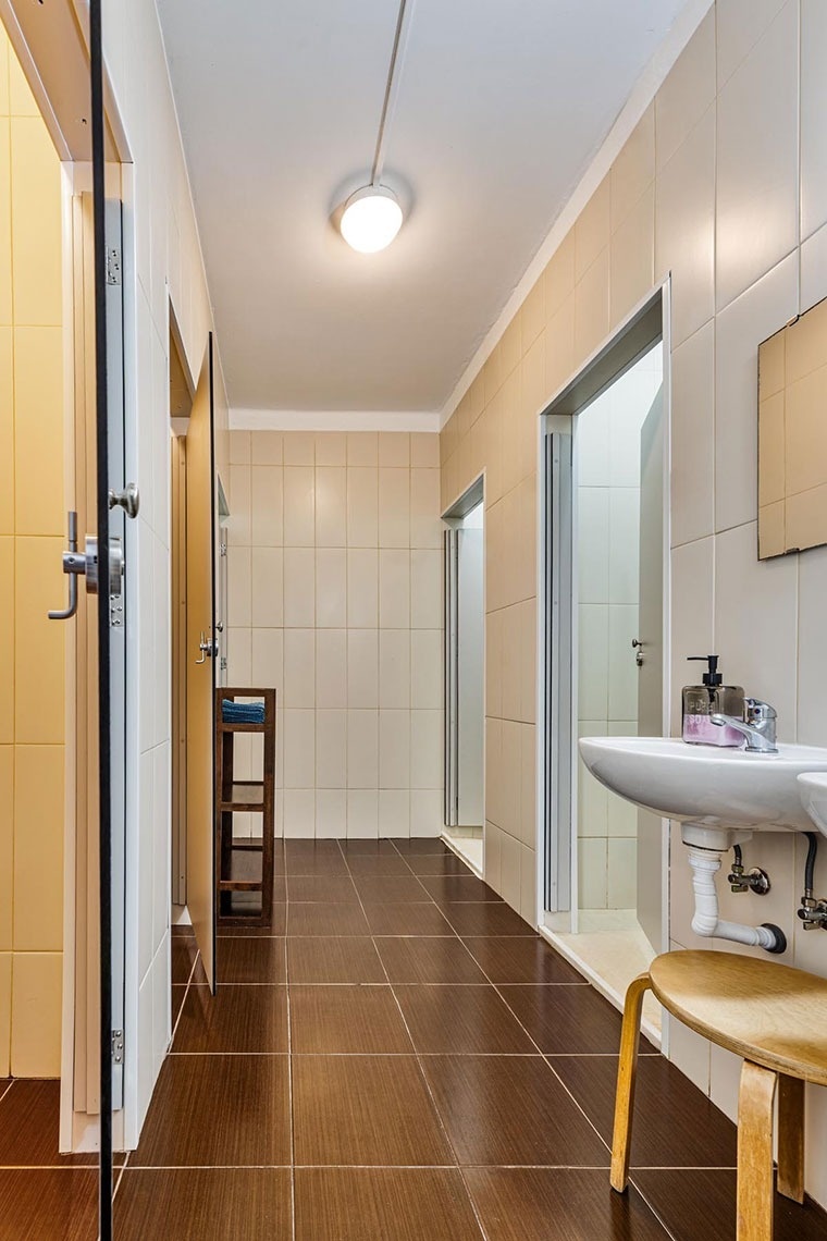 A imagem exibe um corredor de banheiro público com paredes de azulejo claro, chão de azulejo escuro, múltiplas portas para cabines e uma pia com espelho à direita.