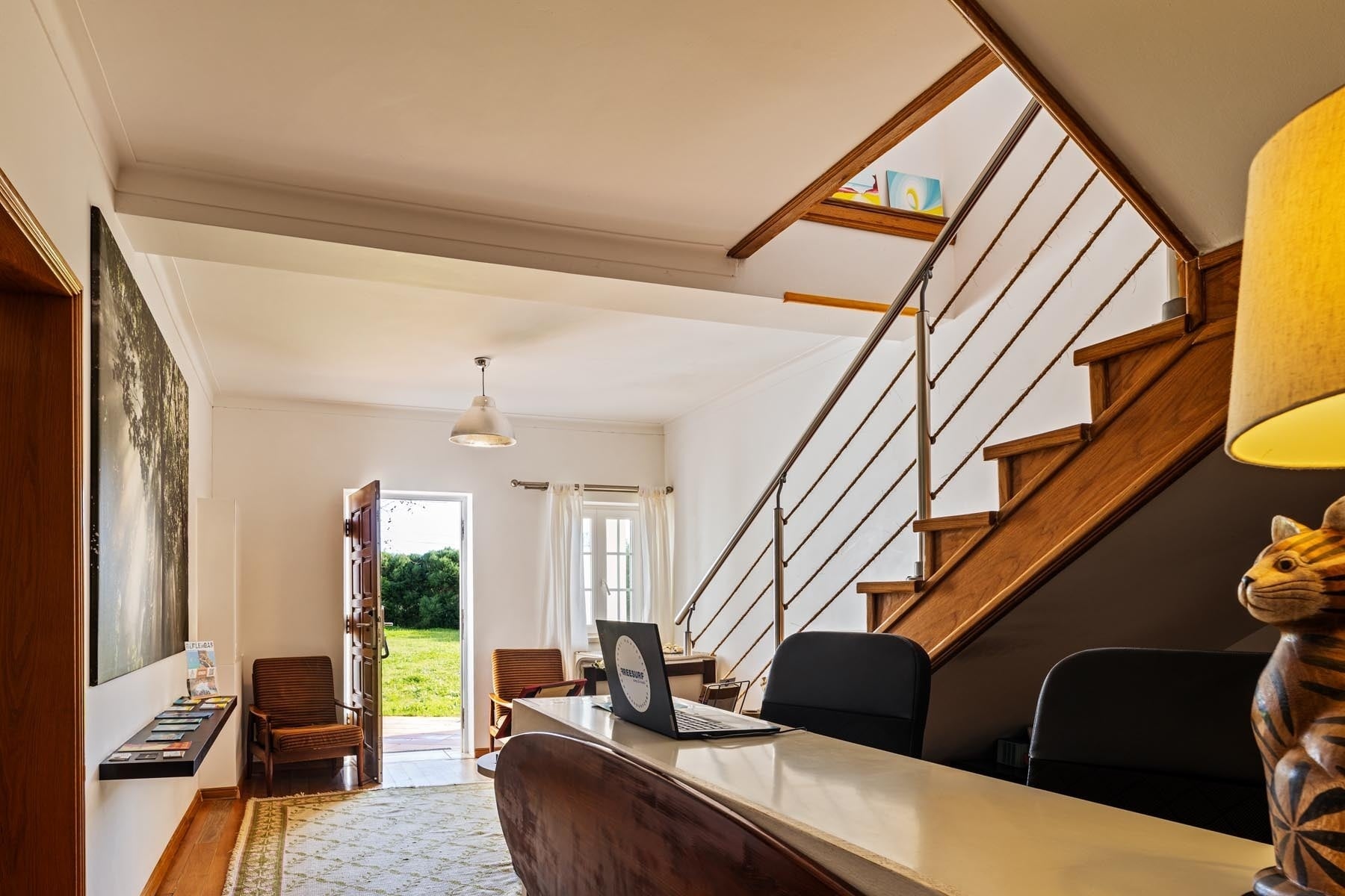 Interior de uma sala luminosa e convidativa com uma escada de madeira à direita, um balcão com um laptop e uma porta aberta para um jardim verde à esquerda.