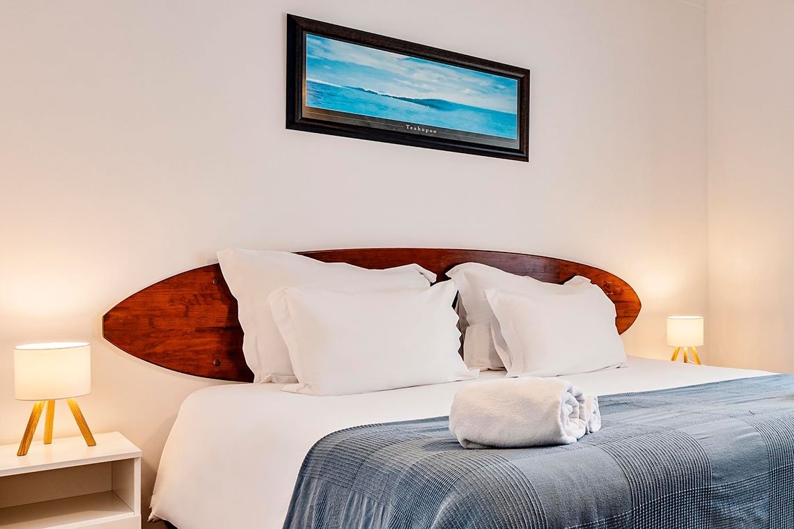 Um quarto de casal arrumado com uma cama com lençóis brancos e manta azul, uma cabeceira de madeira em formato de prancha de surf, duas luminárias de mesa e um quadro de paisagem marinha na parede.