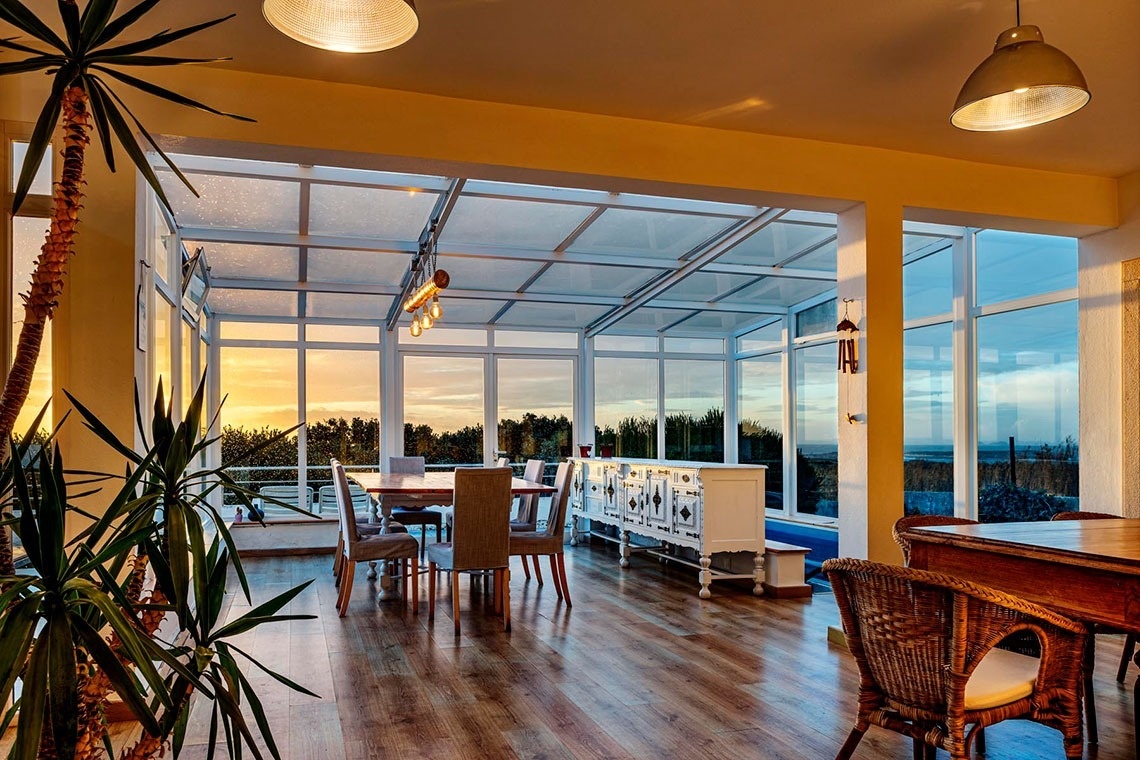 Uma sala de jantar envidraçada e luminosa, com mesa e cadeiras de madeira, um aparador e uma vista panorâmica para o mar e a vegetação, sob um teto de vidro.