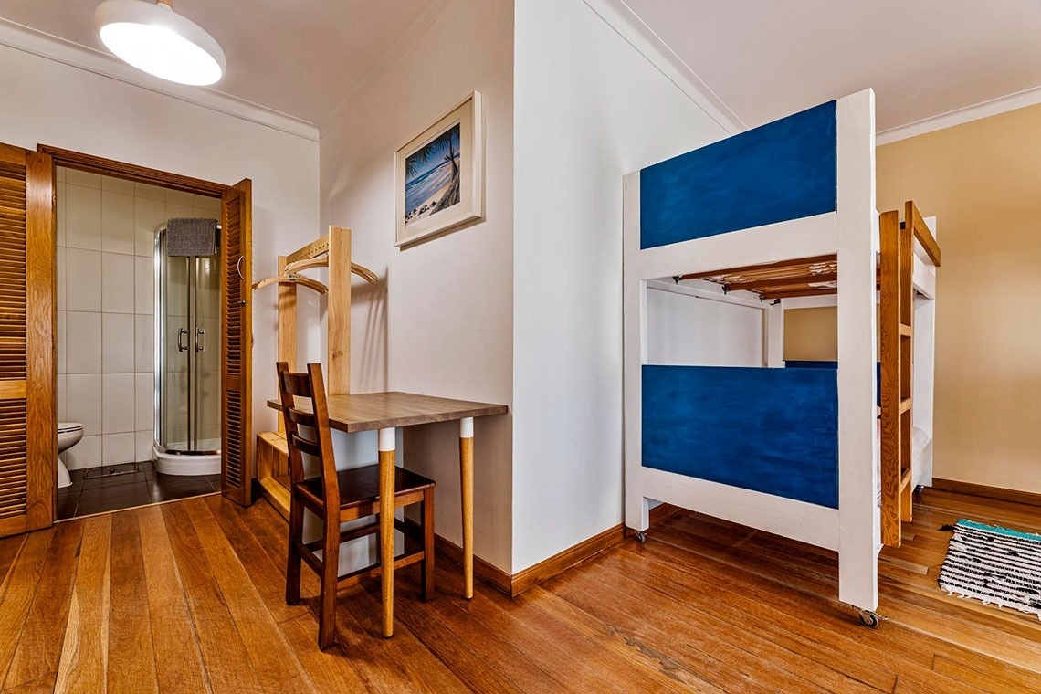 Um quarto funcional com piso de madeira, beliches azul e branco, escrivaninha e banheiro privativo.