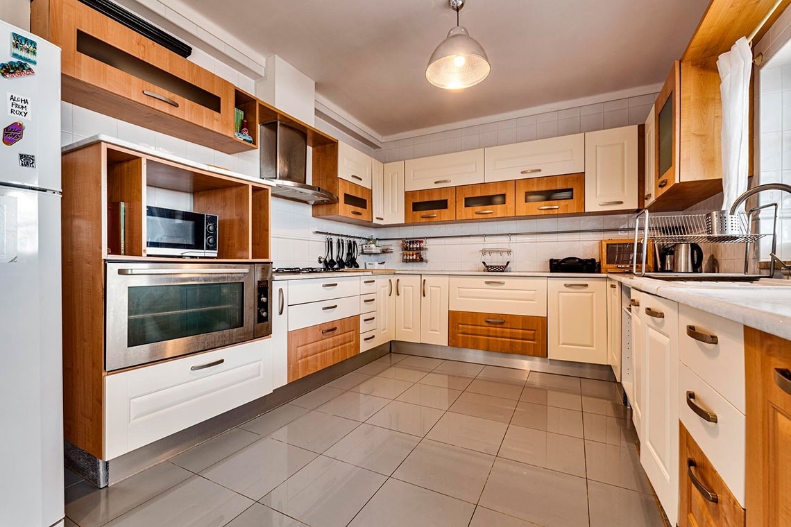 Uma cozinha moderna e espaçosa com armários de madeira clara e creme, chão de azulejos cinzentos e eletrodomésticos integrados.