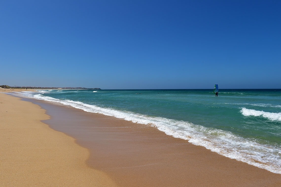 Uma praia com areia dourada e mar azul-turquesa, onde uma pessoa pratica windsurf.