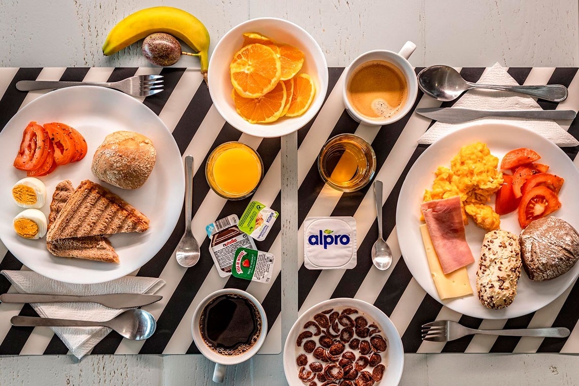 Um café da manhã variado com ovos cozidos, torradas, pão, tomate, suco de laranja e frutas está servido em uma mesa.