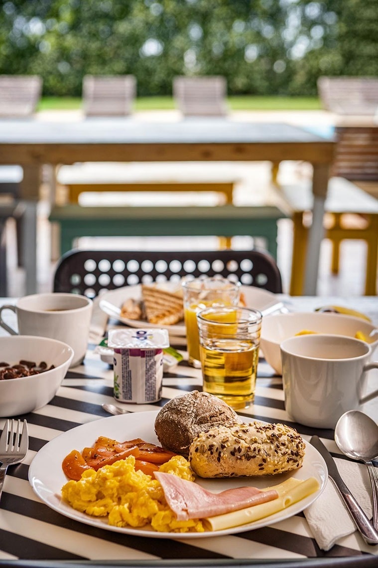Uma mesa farta de café da manhã com ovos mexidos, frios, pães e bebidas é servida em um jogo americano listrado em preto e branco, em um ambiente externo com fundo verde desfocado.