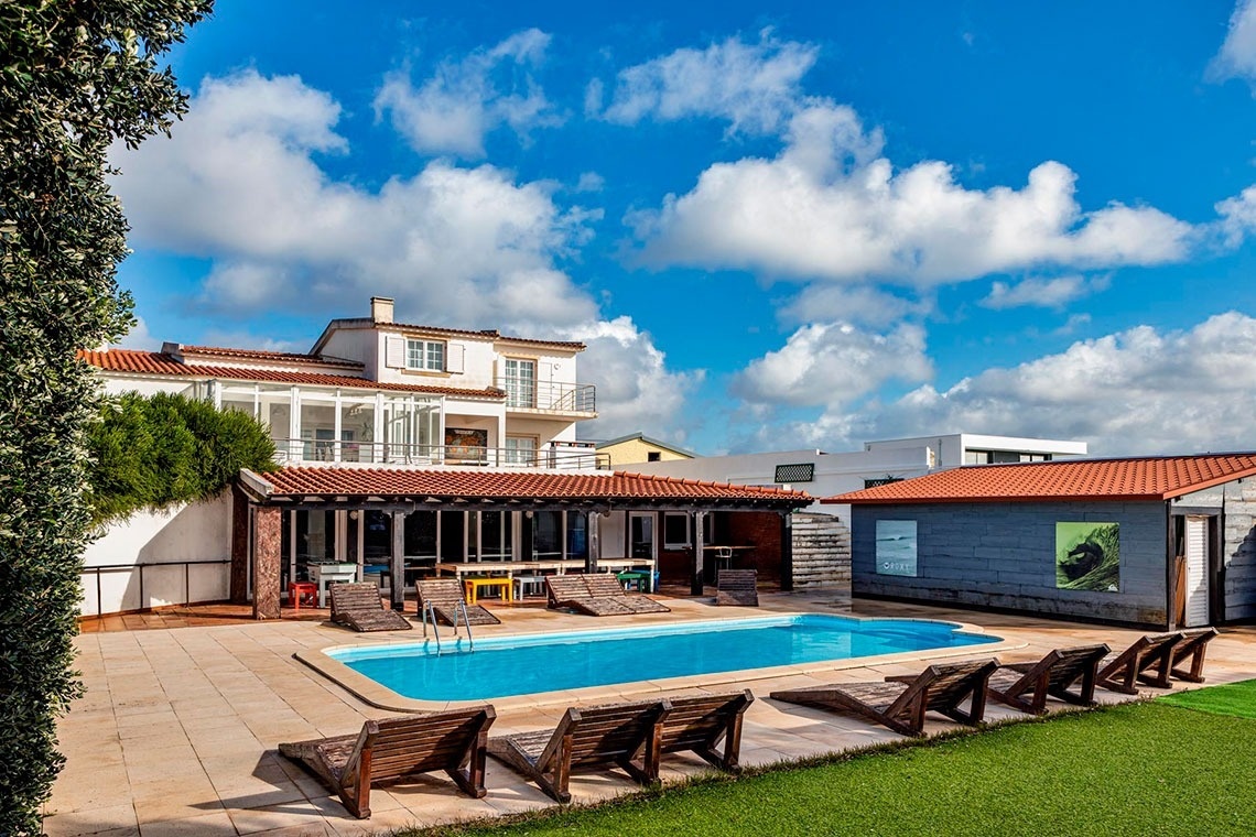 Uma propriedade com uma piscina azul e espreguiçadeiras de madeira é vista num dia ensolarado, com um edifício principal e uma estrutura anexa ao lado, sob um céu azul com nuvens.