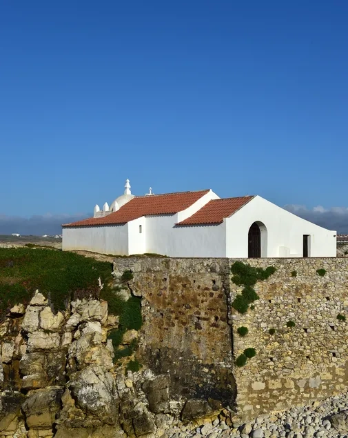 Uma igreja branca de telhado vermelho assenta num penhasco rochoso coberto por vegetação, sob um céu azul límpido.
