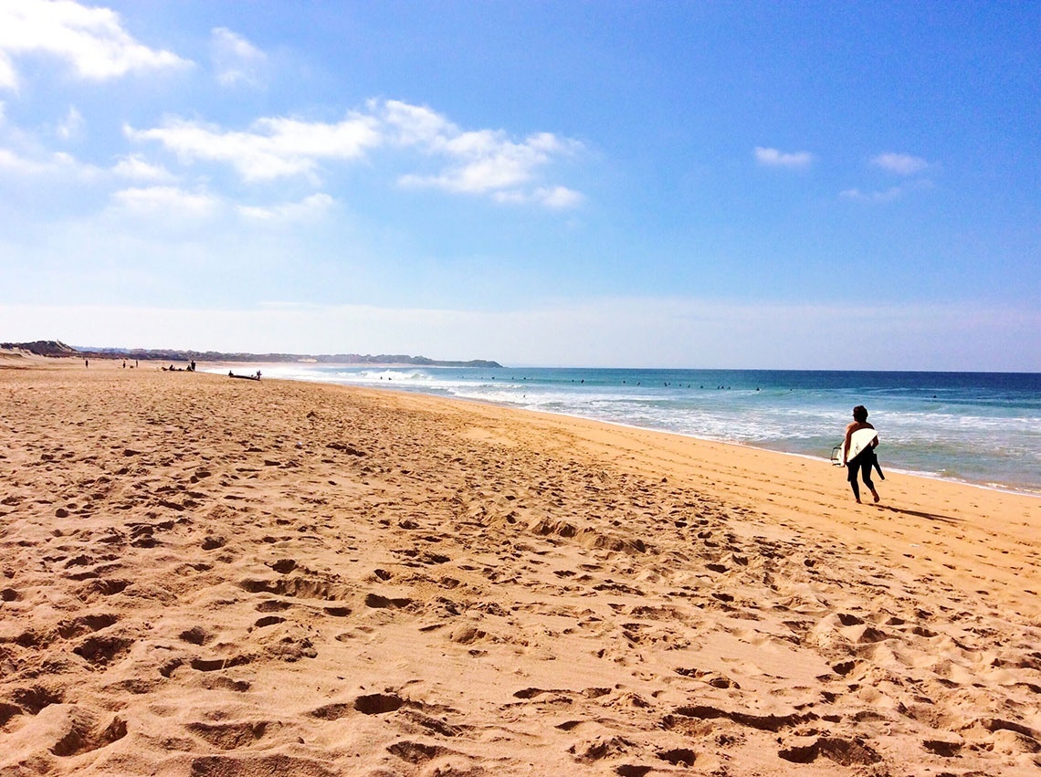 Atardecer en FreesurfCamp: surfista camina por la playa con tabla bajo un cielo azul.