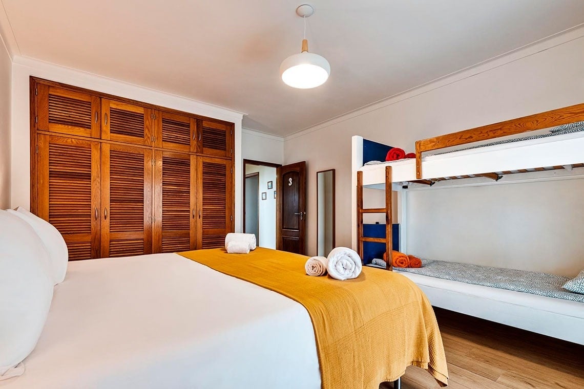 Um quarto com uma cama de casal com cobertor amarelo, um beliche branco e de madeira, e um grande guarda-roupa embutido de madeira.