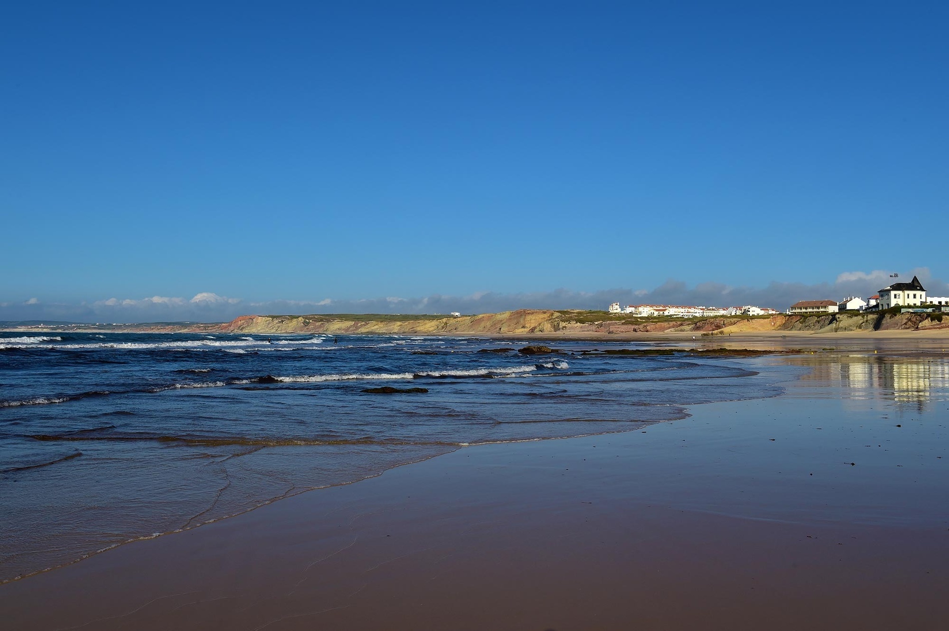 Uma praia extensa com ondas suaves, falésias costeiras e uma vila de casas brancas ao longe, cujos edifícios se refletem na areia molhada sob um céu azul claro.