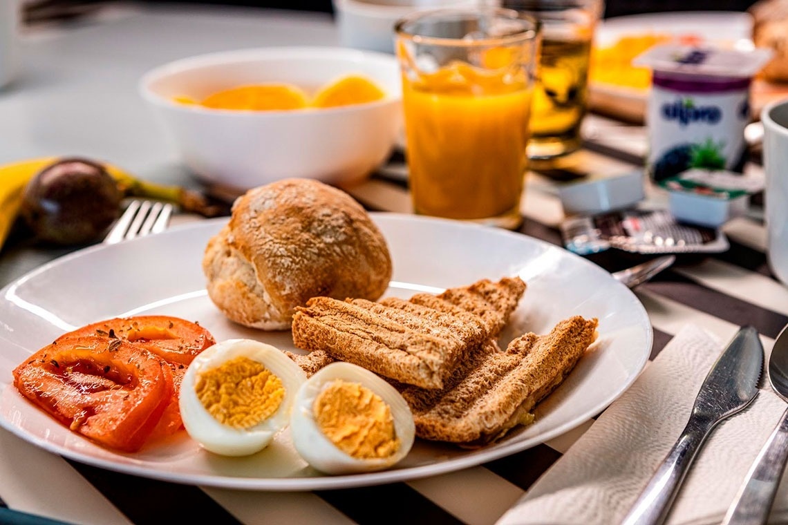 Uma mesa de café da manhã completa é apresentada, com pães, ovos cozidos, tomate, torradas, suco de laranja e frutas.