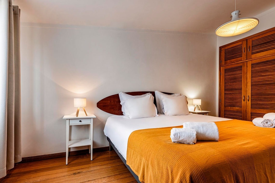 Um quarto claro e acolhedor com uma cama de casal, cabeceira de madeira em forma de prancha de surf e manta laranja, ladeada por mesinhas de cabeceira brancas com abajures, e um guarda-roupa de madeira à direita.