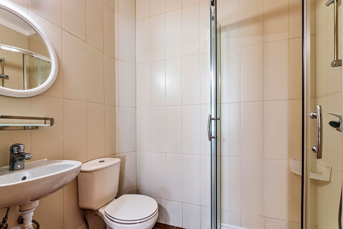 Um banheiro pequeno e funcional com paredes de azulejo bege, pia, vaso sanitário e box de chuveiro de vidro.