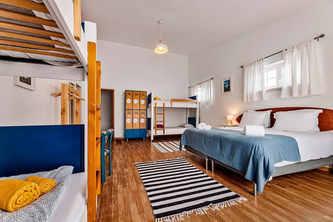 Um quarto luminoso e espaçoso com chão de madeira, paredes brancas, uma cama de casal com roupa de cama azul, vários beliches e armários estilo cacifo.