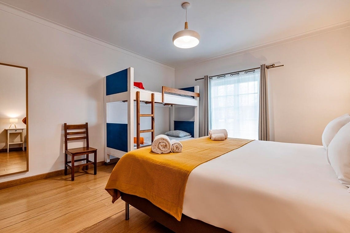 Um quarto de dormir luminoso e organizado apresenta uma cama de casal, um beliche em azul e branco, um espelho de corpo inteiro e uma janela com cortinas claras.