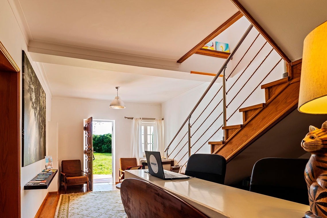 Uma sala interior bem iluminada, possivelmente uma área de recepção, apresenta uma escadaria de madeira, uma secretária com um portátil e uma porta aberta para um jardim verde.