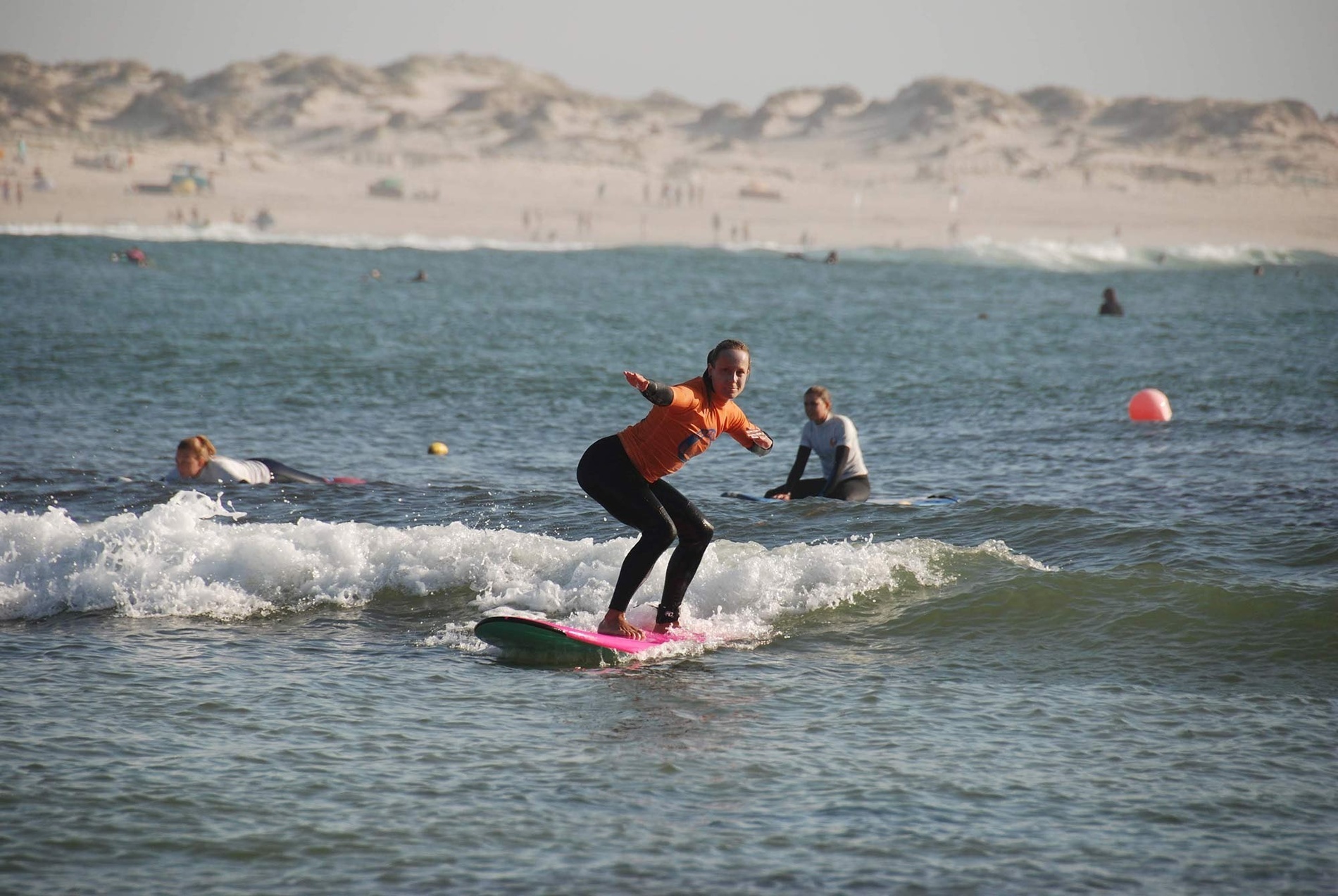 Uma mulher em roupa de surf laranja surfa numa prancha rosa nas ondas do mar, com dunas e outras pessoas ao fundo.