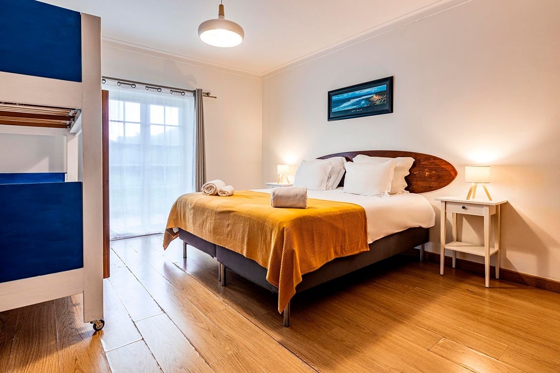 Um quarto de dormir bem iluminado apresenta uma cama feita com roupa de cama branca e uma colcha amarela, piso de madeira, uma mesa de cabeceira com abajur e, à esquerda, parte de um beliche azul.