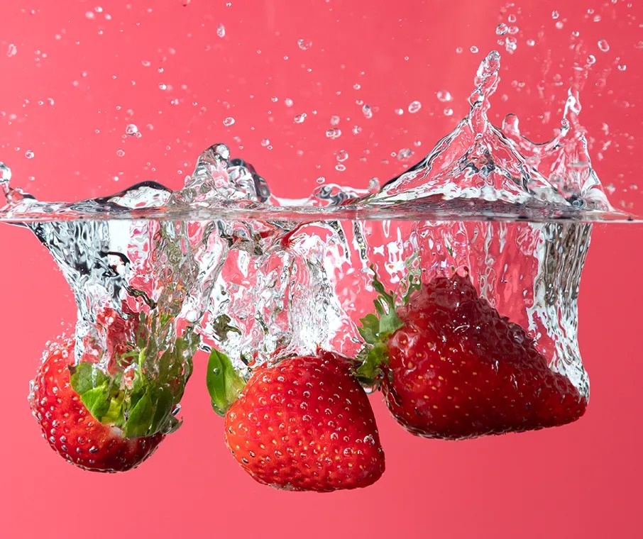 tres fresas caen en el agua sobre un fondo rosa