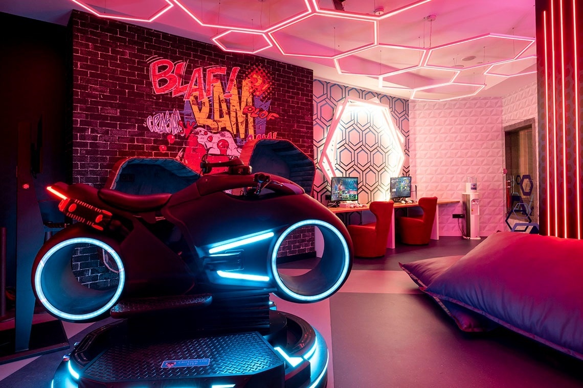 A vibrant, cyberpunk-themed game room features a Tron-like motorcycle simulator, illuminated by red and blue neon lights and hexagonal ceiling fixtures, alongside gaming stations and a graffiti-covered brick wall.