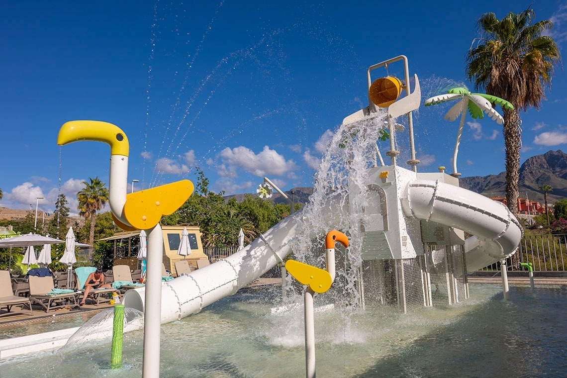 Un parque acuático infantil con un tobogán blanco, chorros de agua, figuras de aves amarillas y palmeras bajo un cielo azul claro.