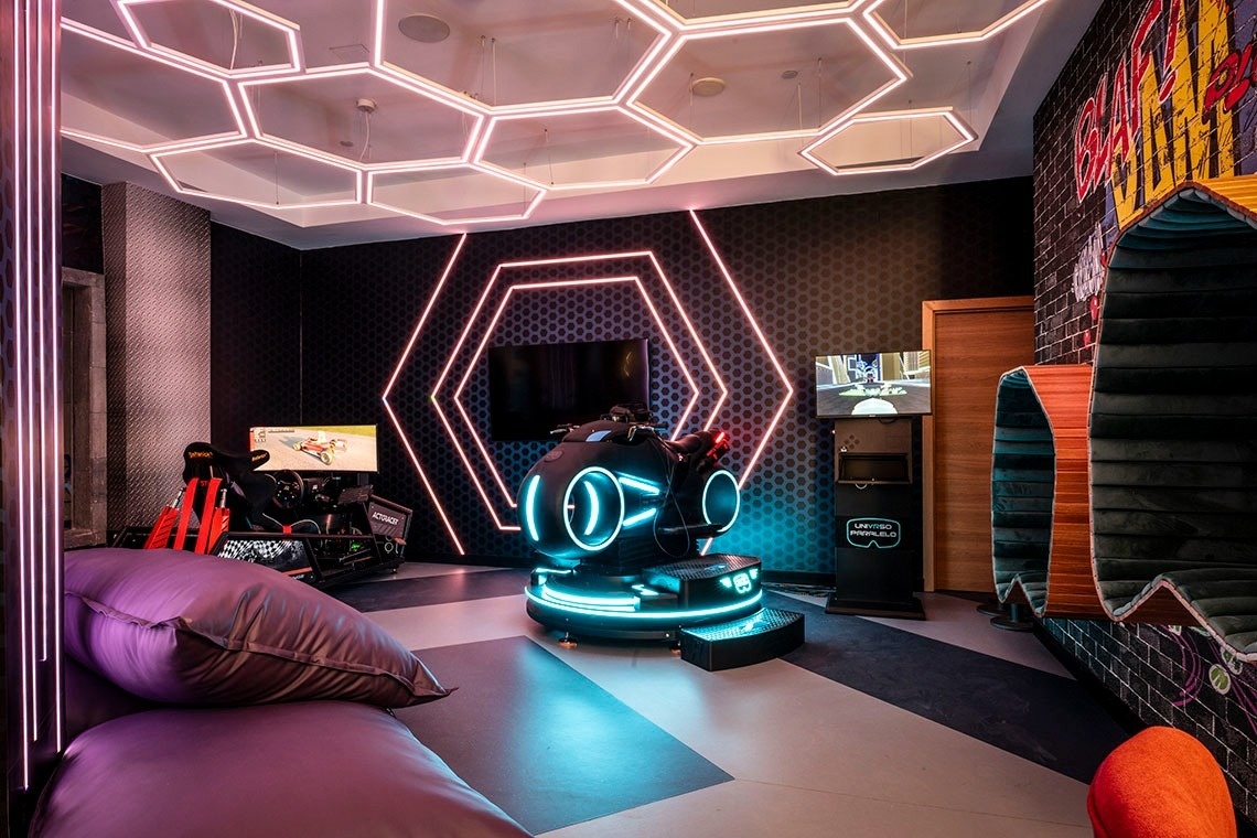 This vibrant, futuristic gaming room is adorned with neon hexagonal lights and features multiple simulators, including a racing rig and a Tron-style motorcycle, alongside a comfortable lounge area.