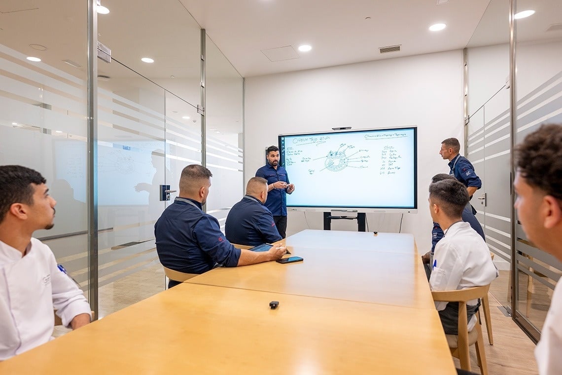 Un instructor culinario da una presentación a un grupo de chefs o estudiantes en una sala de reuniones moderna con una pizarra digital.