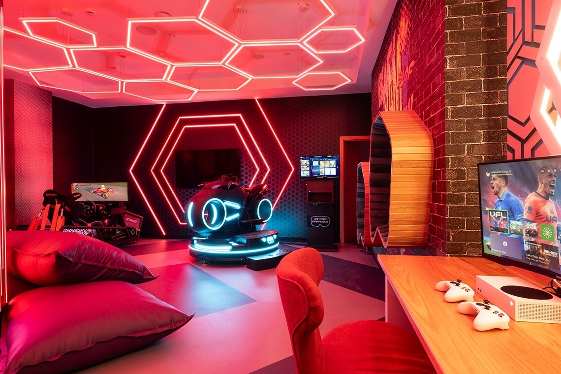 This image captures a vibrant, futuristic gaming room illuminated by red neon hexagonal lights, showcasing various immersive gaming setups including a VR simulator, a racing simulator, and a console gaming station.