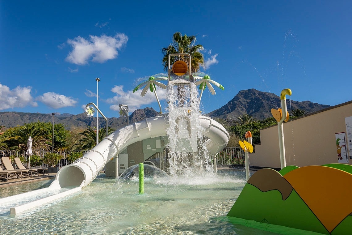 Un vibrante parque acuático con un tobogán blanco, un cubo de agua que se vuelca y diversas fuentes de chorros, se sitúa bajo un cielo azul con montañas y palmeras al fondo.