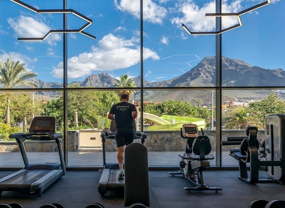 A person runs on a treadmill in a modern gym with large windows offering a panoramic view of mountains, palm trees, and a water slide under a bright blue sky.