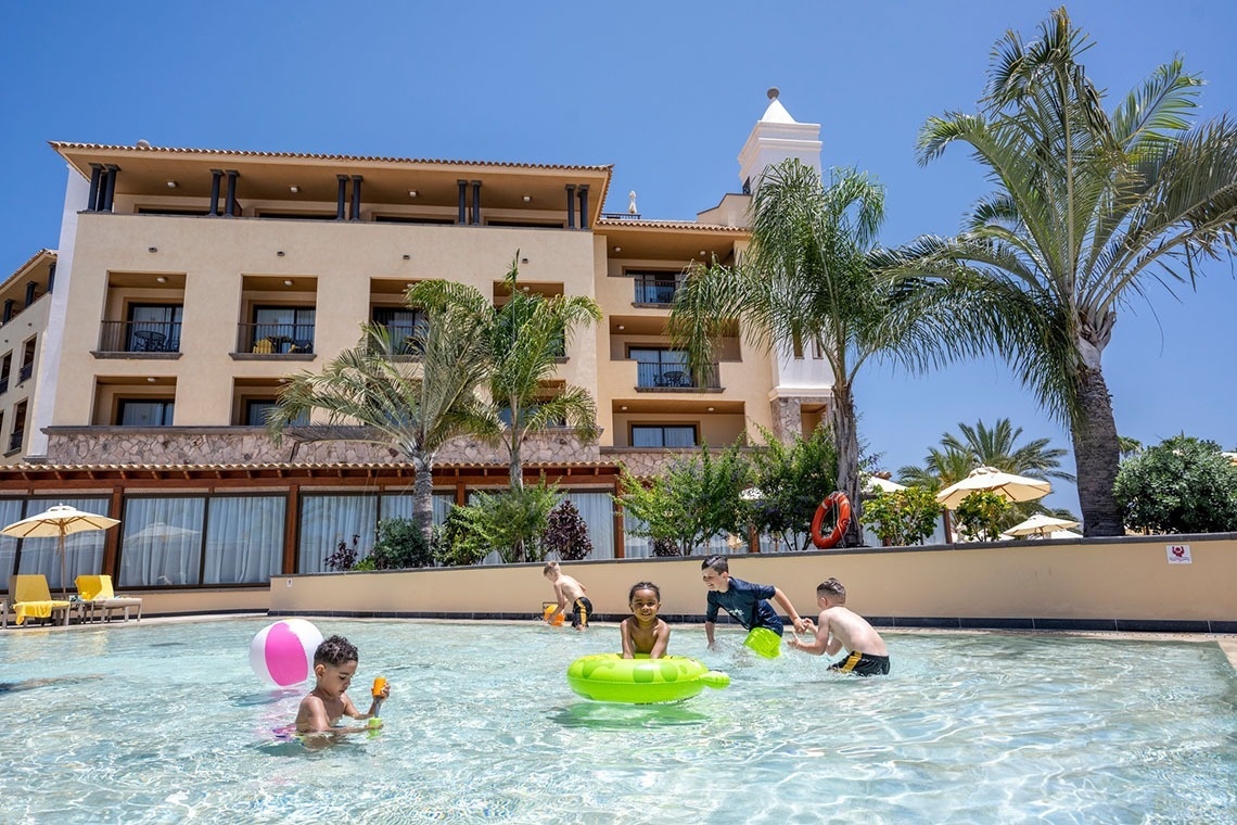 Varios niños se divierten en una piscina de un resort, con un edificio y palmeras al fondo, bajo un cielo azul.