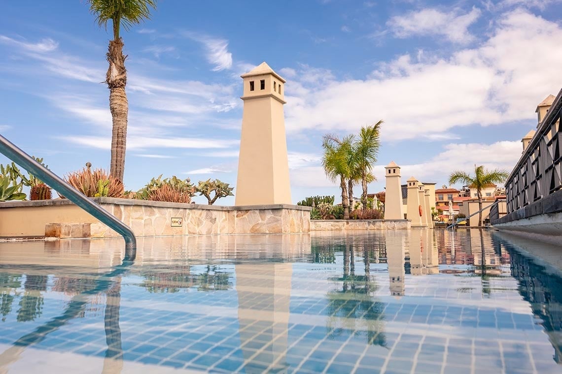 A tranquil infinity pool with clear blue water and reflections of palm trees and distinctive chimney-like structures under a bright sky.