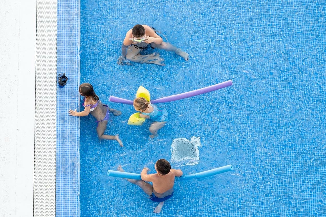 Una vista aérea muestra a varias personas, incluyendo adultos y niños, disfrutando de una piscina de baldosas azules con algunos usando flotadores.