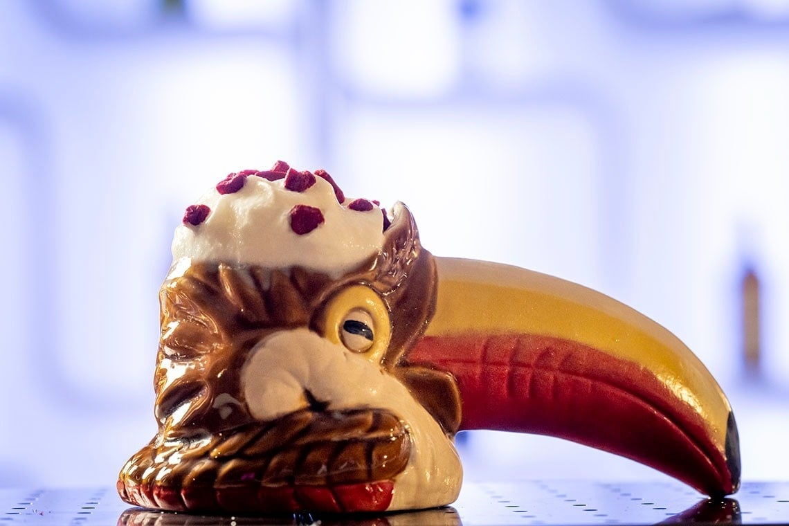 Una bebida cremosa blanca con trocitos rojos se sirve en una taza tiki con forma de tucán, de colores vibrantes, sobre un fondo luminoso y desenfocado.