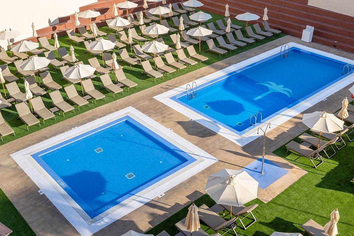La imagen muestra una vista aérea de un complejo turístico con dos piscinas, rodeadas de numerosas tumbonas y sombrillas en un área verde y pavimentada.