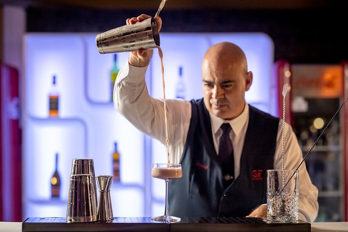 Un barman calvo vierte una bebida rosada de una coctelera en una copa elegante en un bar.