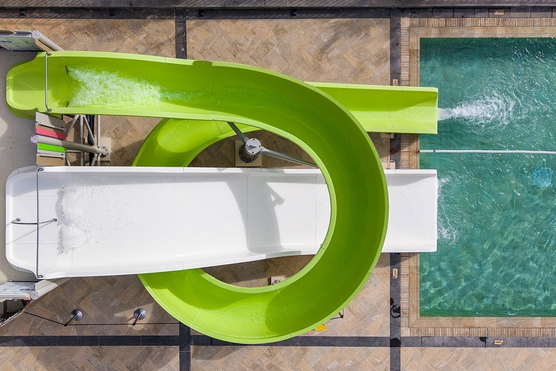 Una vista aérea muestra dos toboganes de agua, uno verde y uno blanco, desembocando en una piscina de azulejos.