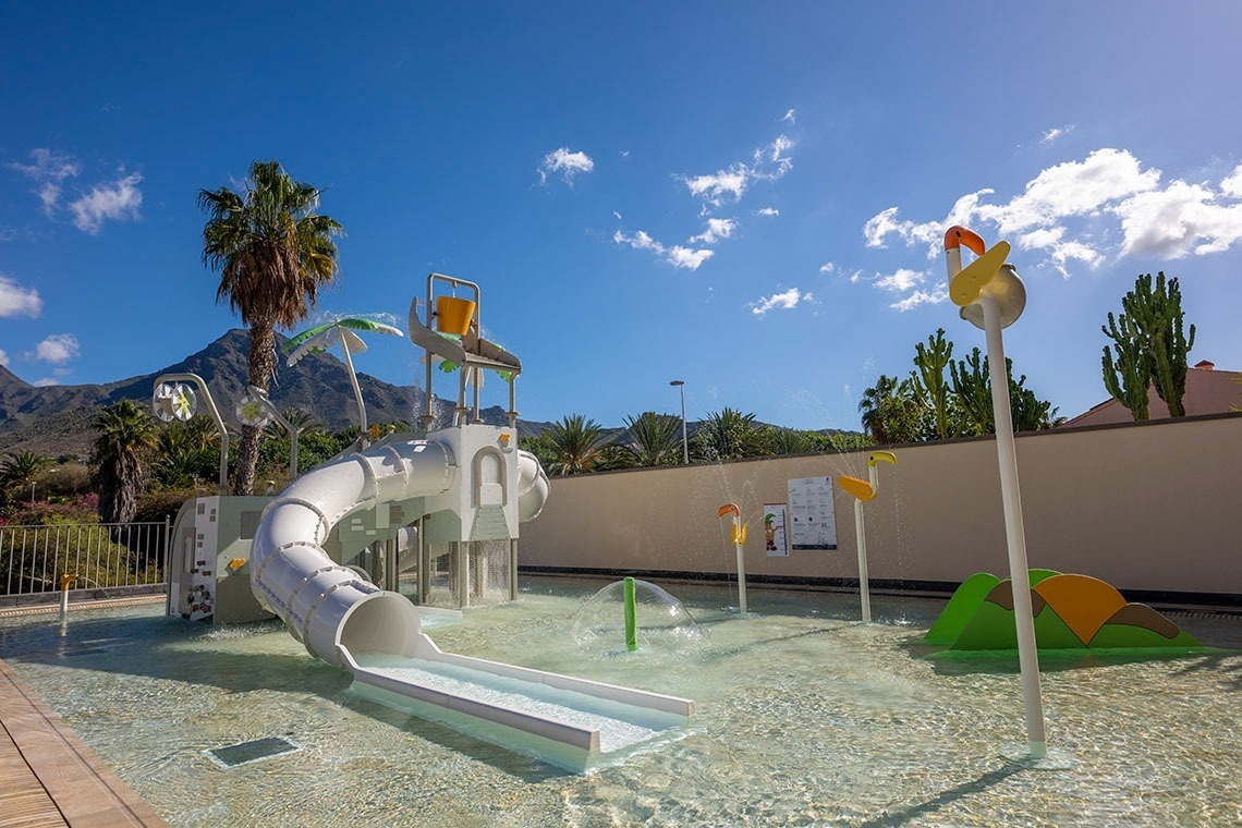 Un parque acuático infantil con tobogán y juegos de agua, rodeado de palmeras y montañas bajo un cielo azul.