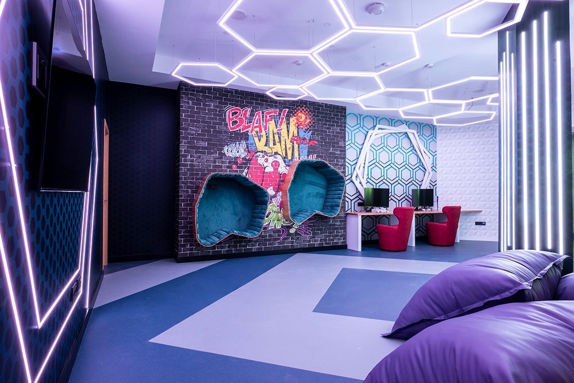 This vibrant and modern lounge space features hexagonal ceiling lights, neon strip lighting, bold graffiti art on a brick wall, and various seating options including teal pod chairs, red armchairs, and purple beanbags.