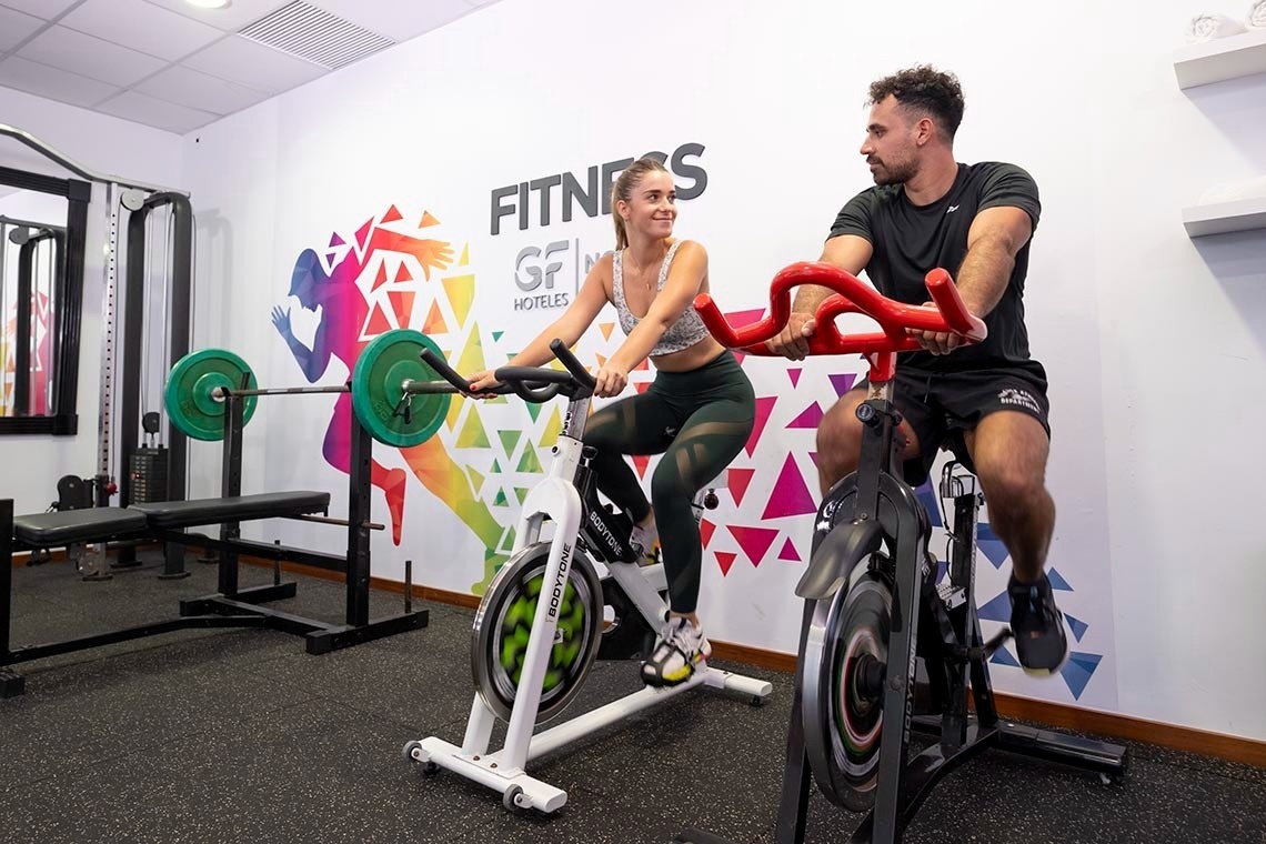 A man and a woman are smiling and looking at each other while exercising on stationary bikes in a brightly lit gym.