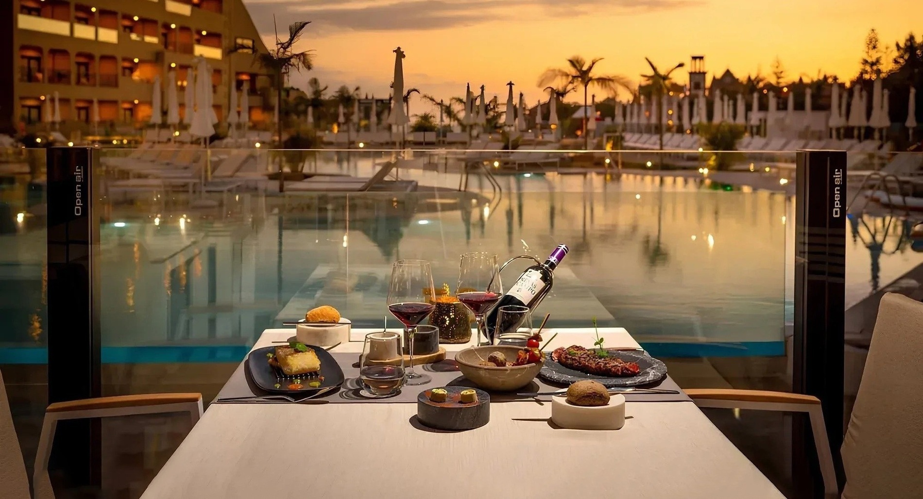 una mesa con comida y una botella de vino frente a una piscina al atardecer