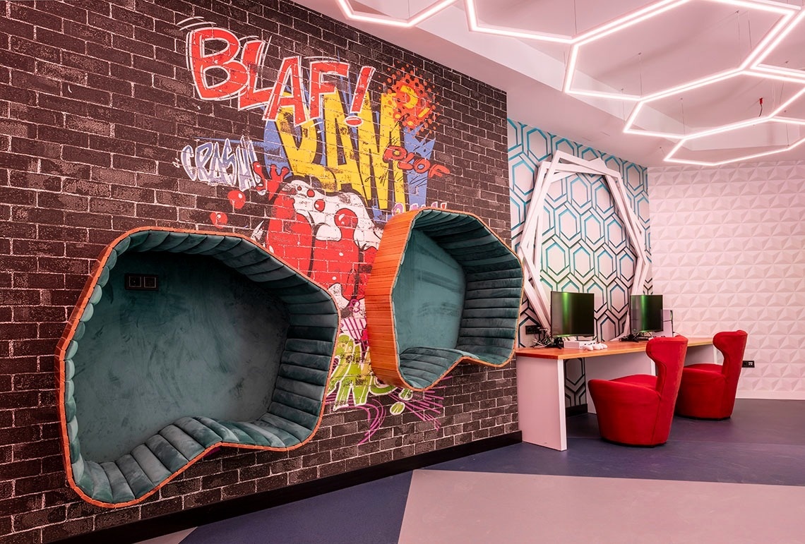This lively entertainment space showcases two plush, pod-like seats embedded in a graffiti-covered brick wall, alongside a dual-monitor gaming station and striking hexagonal ceiling lights.