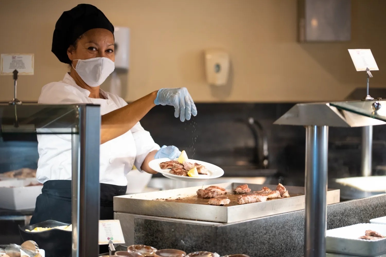 una cocinera con una máscara y guantes añade sal a un plato de carne