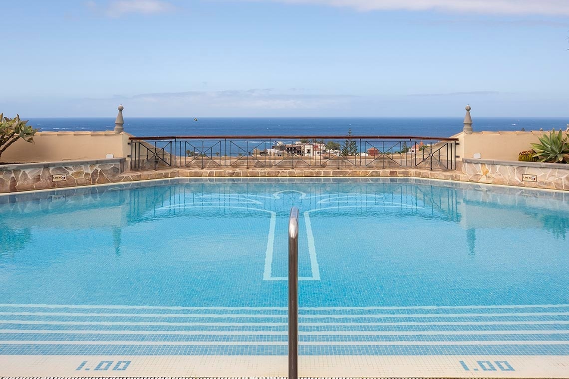 An elevated, blue-tiled swimming pool featuring a palm tree mosaic offers a panoramic view of the vast blue ocean and distant coastline under a clear sky.