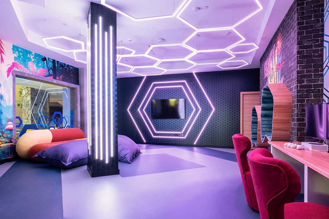 This vibrant, futuristic-themed room features striking hexagonal neon lights on the ceiling and a wall, a large television, a gaming setup with red chairs, and colorful lounge seating, all illuminated with a purple glow.
