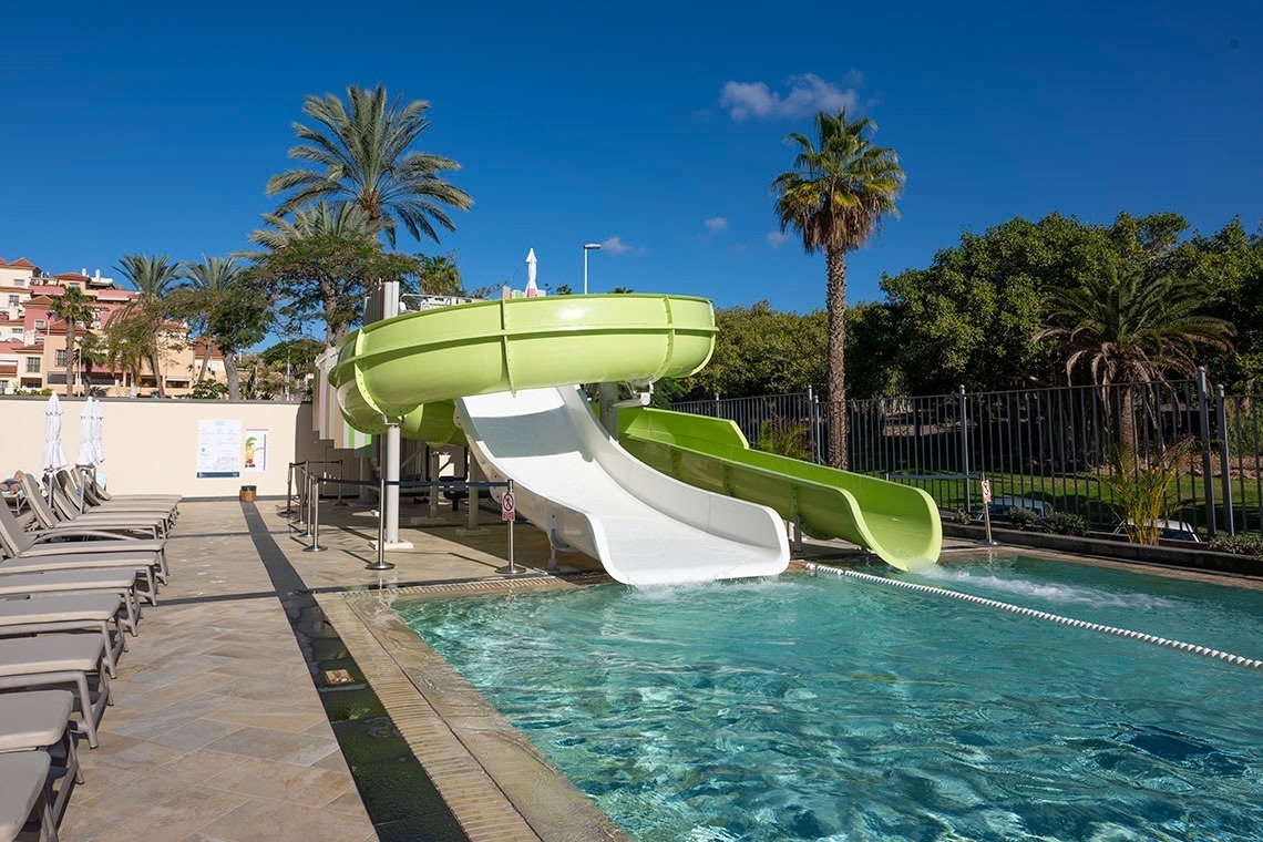 Una piscina con toboganes de agua, rodeada de tumbonas y palmeras, bajo un cielo azul claro.